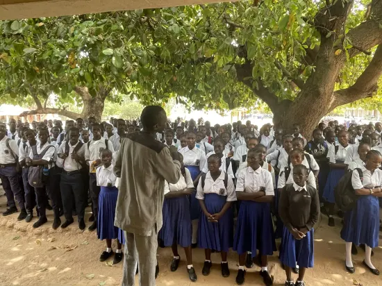 Secondary school assembly gathered outdoors under a large tree, The Gambia, 2022